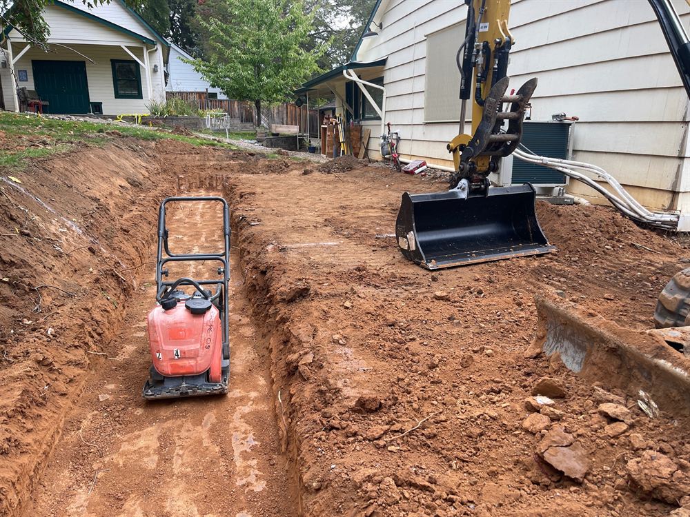 Utility trench excavation beside a home with a plate compactor and mini excavator